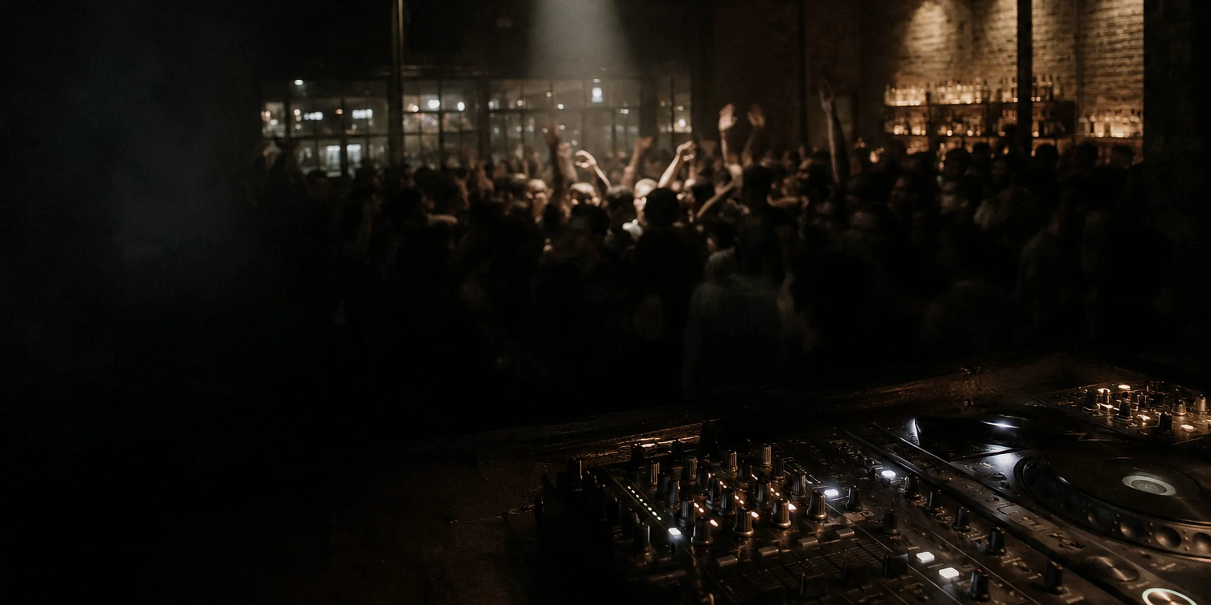 View from the DJ booth across a mixer and CDJ to a crowd with hands raised under a single spotlight, backbar glowing along the far wall