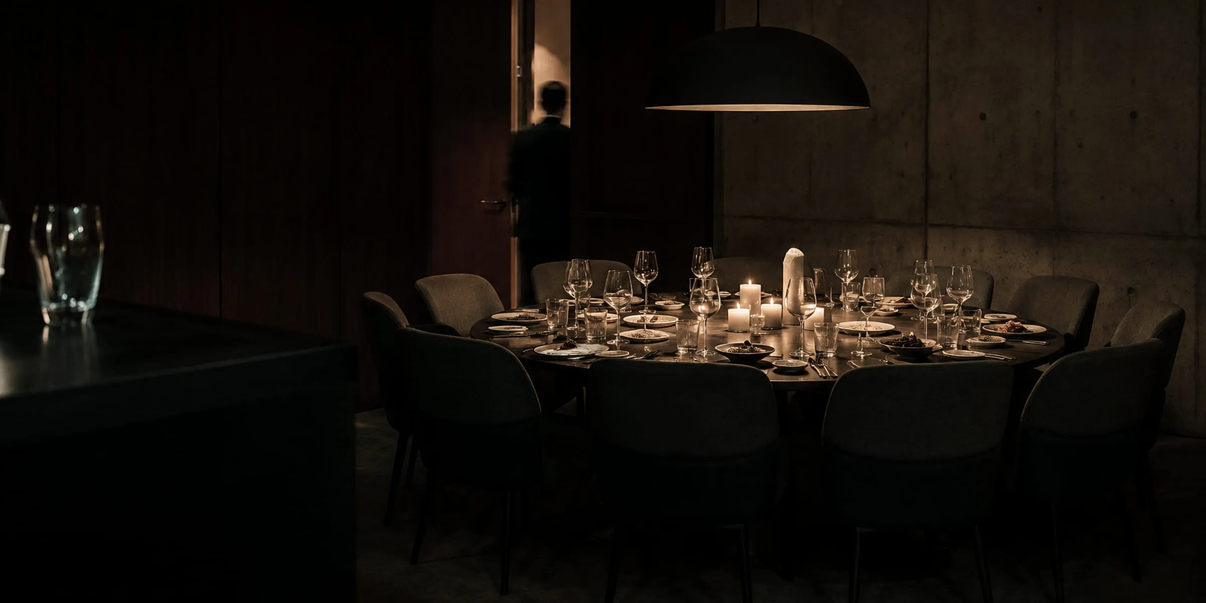 Private dining room lit by one wide pendant over a round table set with candles and glassware, concrete and dark timber walls