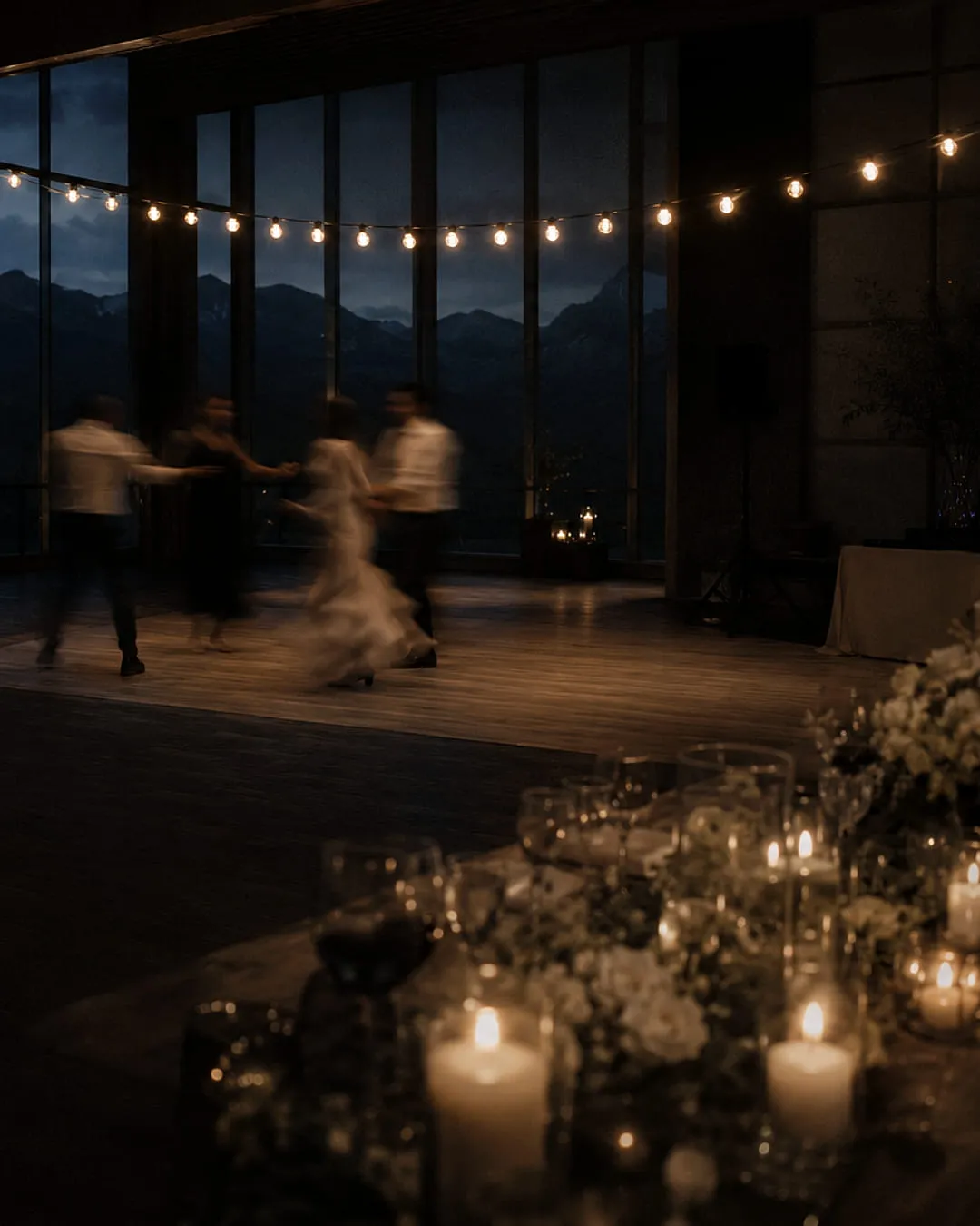 Reception room at dusk with a wall of glass onto mountains, string lights overhead, guests dancing, candles and white florals along the foreground table