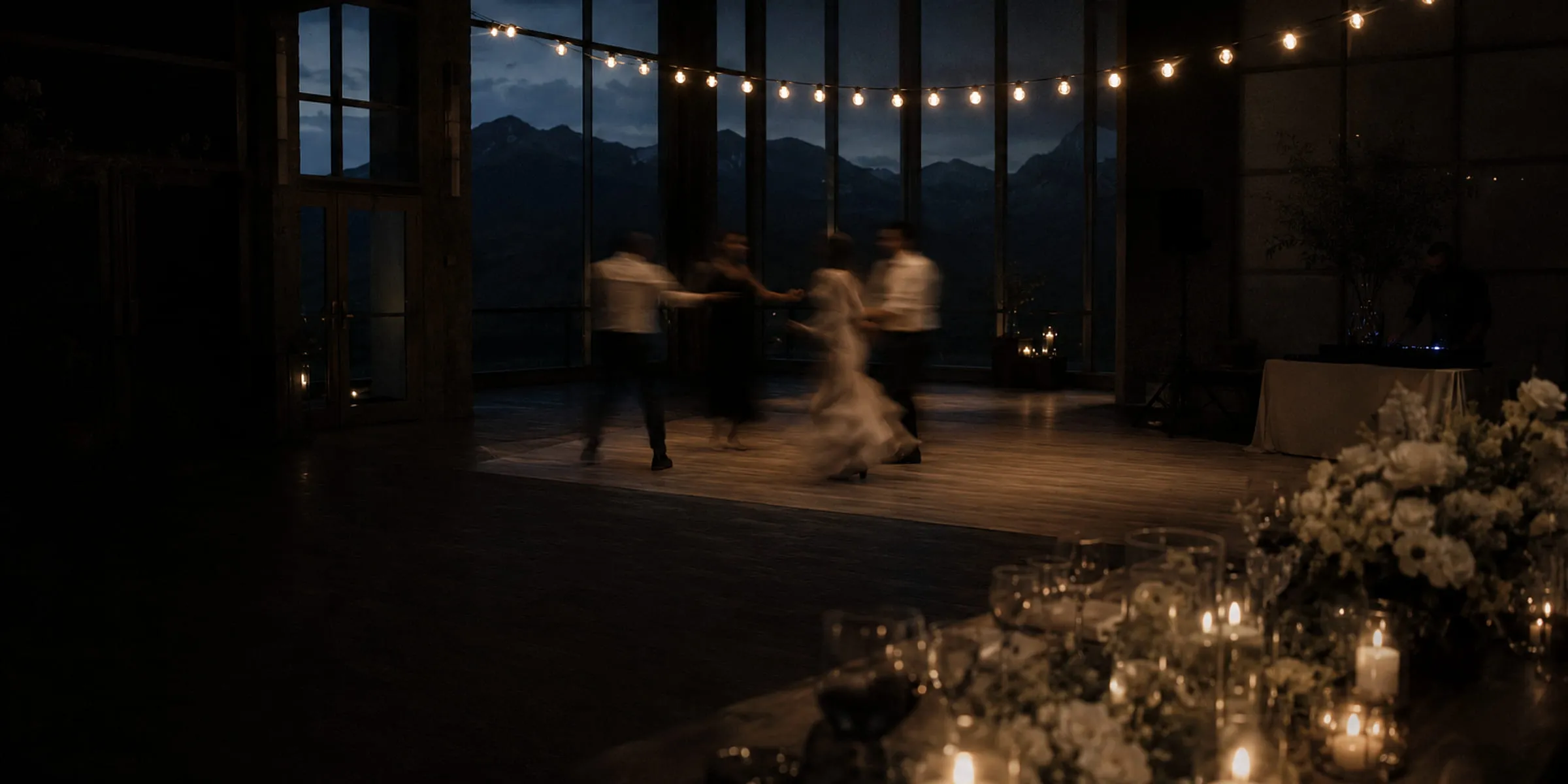 Reception room at dusk with a wall of glass onto mountains, string lights overhead, guests dancing, candles and white florals along the foreground table